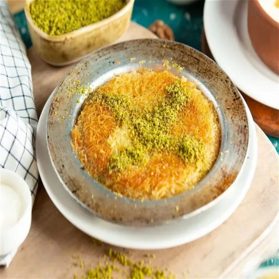 Golden kunefe dessert in a metal pan, topped with green pistachios, on a white plate.